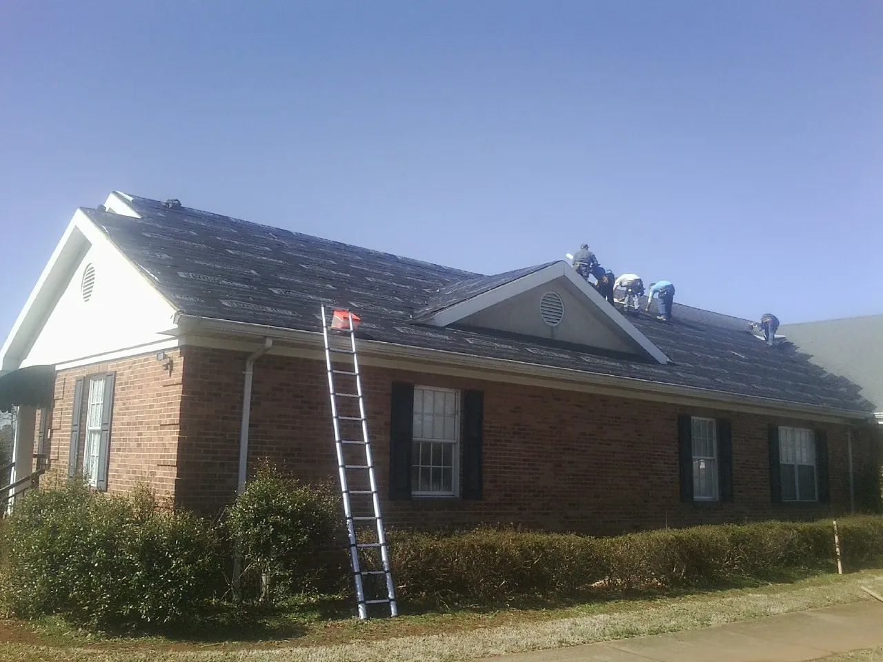 Crew tearing shingles off a brick ranch roof with a ladder set — Emergency Roof Repair work by our crew in Danese