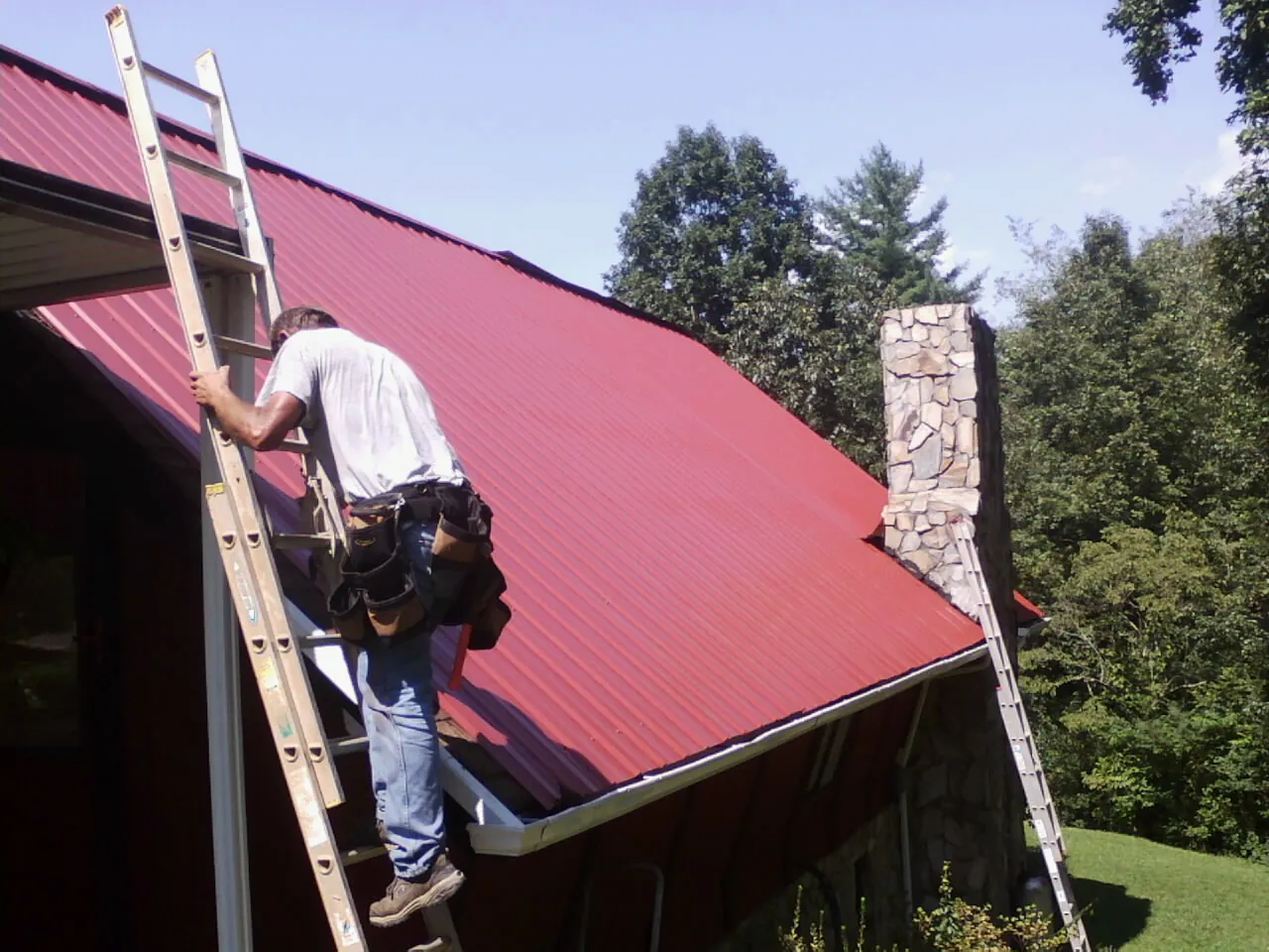 Roofer on a ladder beside a red metal roof and stone chimney — Soffit & Fascia Repair by Roofing Brothers in Danese